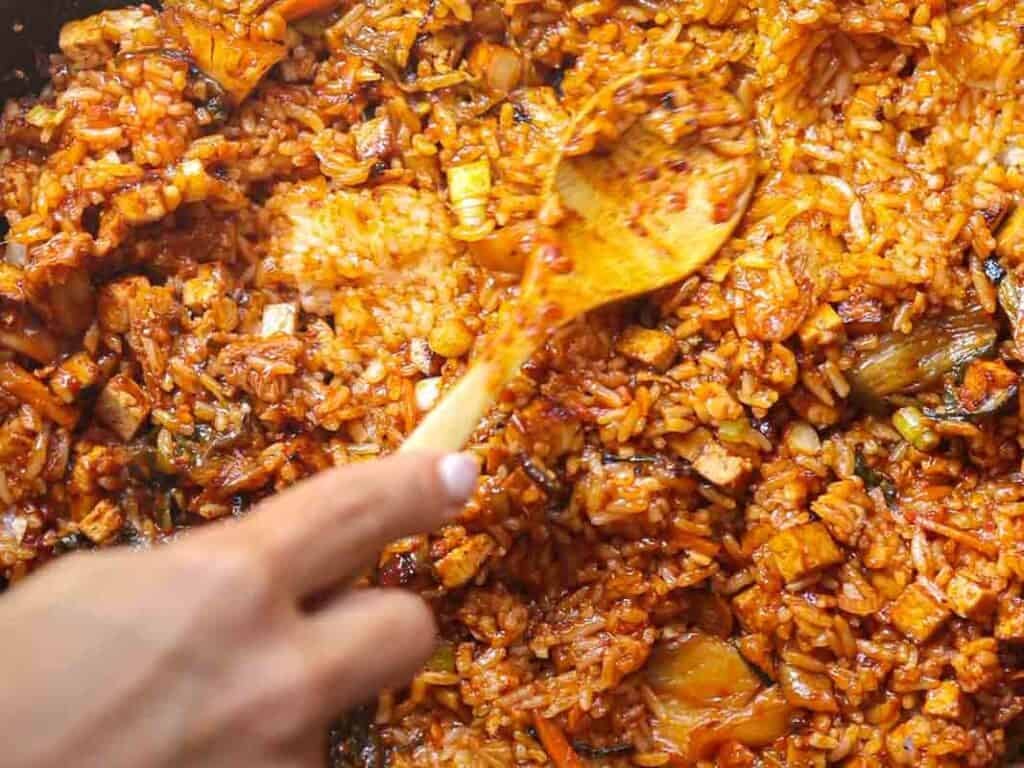 A close-up of a hand stirring kimchi fried rice in a pan with a wooden spoon. The dish includes rice, pieces of kimchi, and diced vegetables, all cooked with a reddish seasoning.